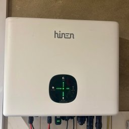 Biały inwerter marki Hinen zamontowany na ścianie z widocznymi kablami i wskaźnikami LED, prezentujący instalację fotowoltaiczną wewnątrz budynku.