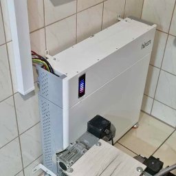 Biały magazyn energii Hinen w pomieszczeniu z kafelkami, obok skrzynka z bezpiecznikami. Widoczne kable i elementy instalacji. Ujęcie z dołu, podkreślające techniczny charakter.