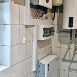 IQDOM24 SPÓŁKA Z OGRANICZONĄ ODPOWIEDZIALNOŚCIĄ - Instalacja urządzeń w kotłowni: biała obudowa inwertera z okablowaniem, rozdzielnica, zbiornik buforowy i piec z palnikiem, na tle wykafelkowanej ściany.