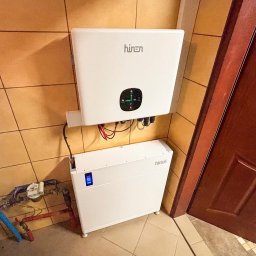 IQDOM24 SPÓŁKA Z OGRANICZONĄ ODPOWIEDZIALNOŚCIĄ - Biały, dwuczęściowy magazyn energii Hinen zamontowany na kafelkowej ścianie, z widocznymi przewodami i elementami instalacji wodnej w tle, obok brązowych drzwi.