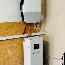 IQDOM24 SPÓŁKA Z OGRANICZONĄ ODPOWIEDZIALNOŚCIĄ - Biały inwerter z podłączonymi kablami i biały akumulator SLUXER na tle ściany z żółtą lamperią. Widoczne przewody i elementy instalacji.