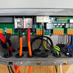 IQDOM24 SPÓŁKA Z OGRANICZONĄ ODPOWIEDZIALNOŚCIĄ - Wnętrze skrzynki elektrycznej z licznymi przewodami o różnych kolorach, podłączonymi do zielonej płytki drukowanej. Widoczne złącza, zaciski i elementy elektroniczne.