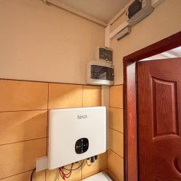 IQDOM24 SPÓŁKA Z OGRANICZONĄ ODPOWIEDZIALNOŚCIĄ - Biały falownik marki Hinen zamontowany na kafelkowanej ścianie, obok brązowych drzwi i skrzynki elektrycznej. Widoczne przewody.