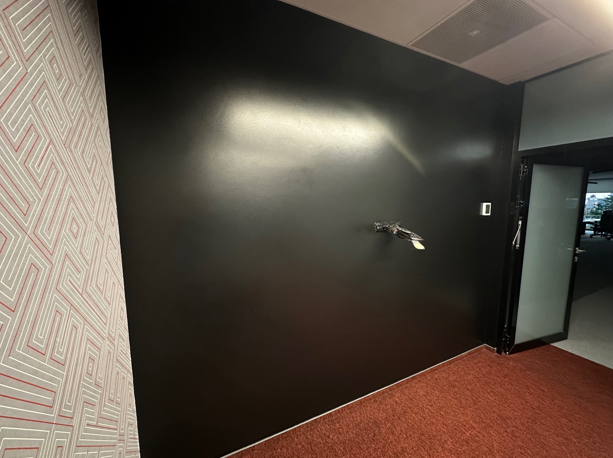 Ciemna, gładka ściana po malowaniu w biurze, widoczne gniazdko elektryczne i fragment wzorzystej tapety obok. W tle przeszklone drzwi do innego pomieszczenia.