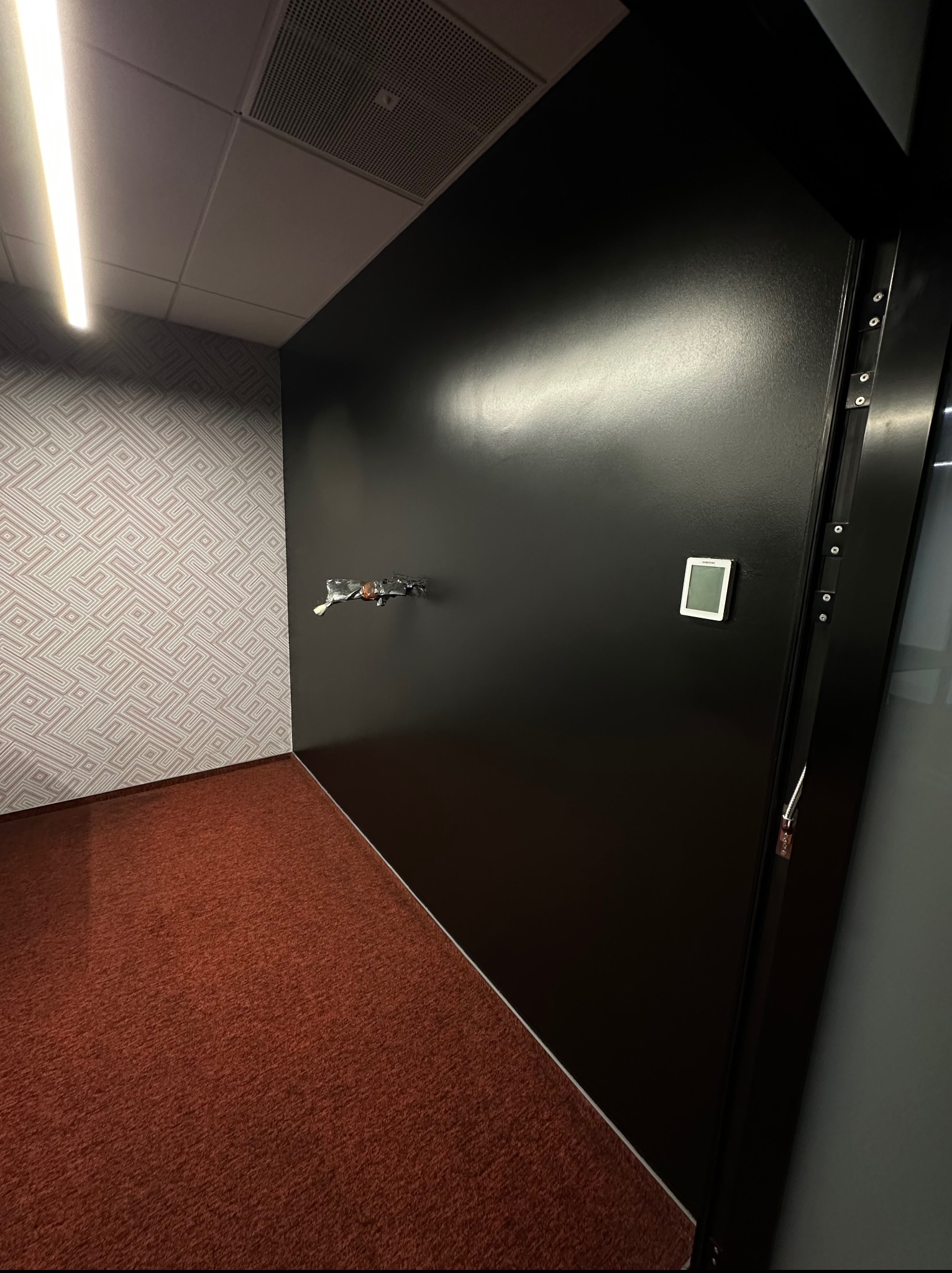 Wnętrze biura po remoncie: gładka, czarna ściana z czujnikiem temperatury, wzorzysta tapeta i czerwona wykładzina. Fragment instalacji elektrycznej wystaje ze ściany.