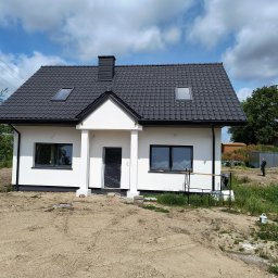 Andrzej Szewczyk usługi remontowo-budowlane - Biały dom jednorodzinny z ciemną dachówką i oknami, widok z zewnątrz. Świeżo otynkowana elewacja, teren wokół w trakcie zagospodarowania. W tle budynek gospodarczy.