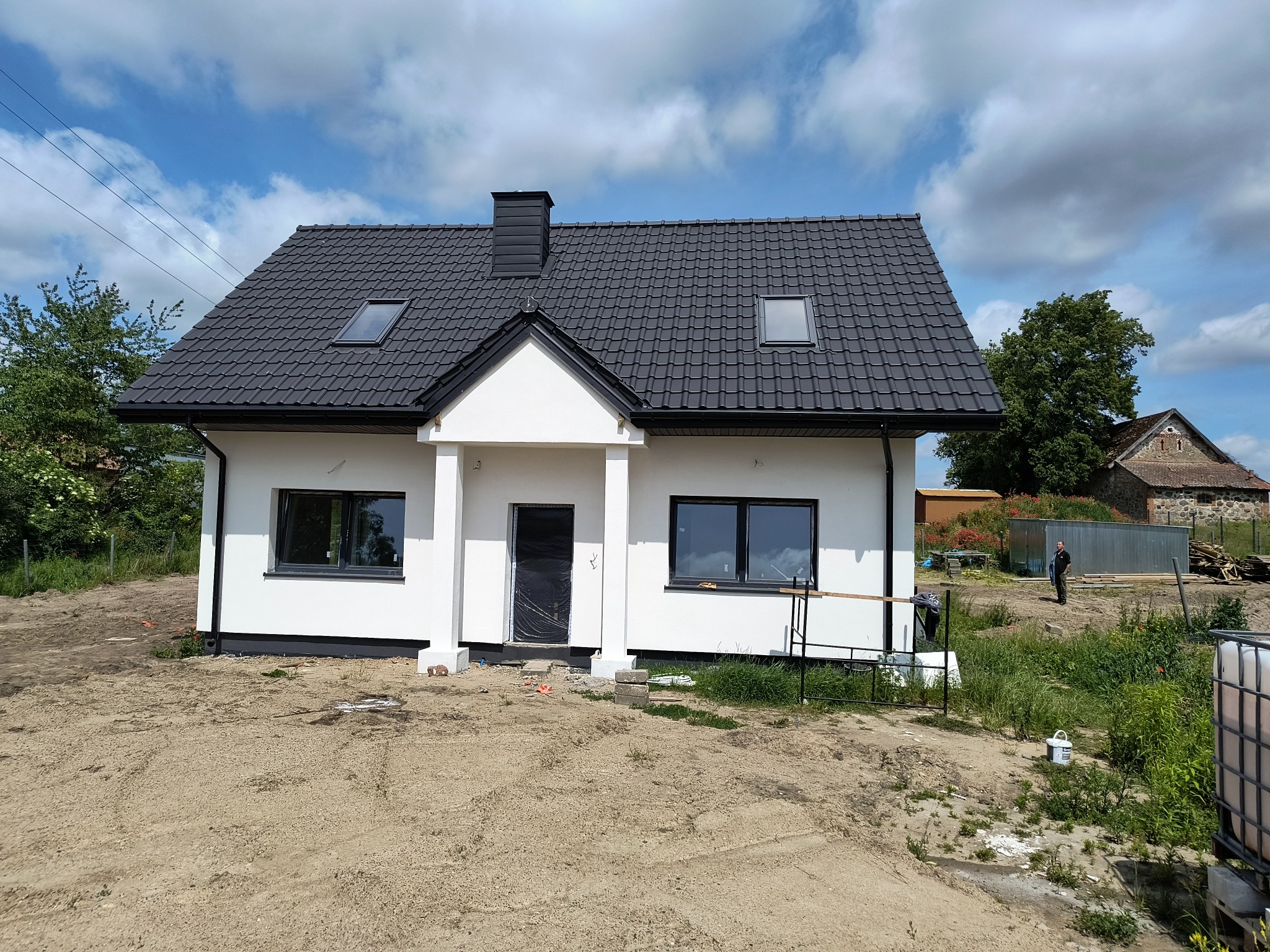 Biały dom jednorodzinny z ciemną dachówką i oknami, widok z zewnątrz. Świeżo otynkowana elewacja, teren wokół w trakcie zagospodarowania. W tle budynek gospodarczy.