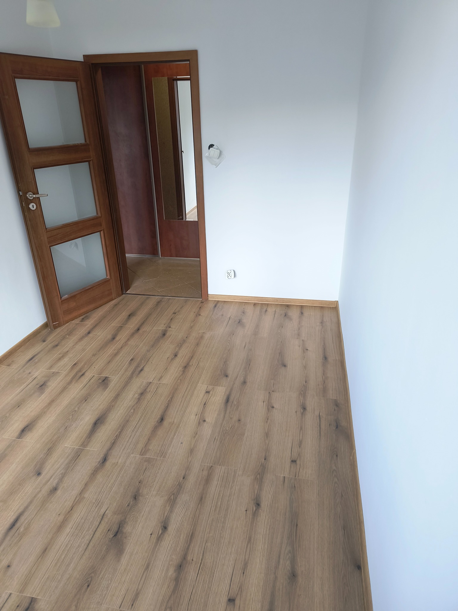 Pusty pokój z drewnianą podłogą, białymi ścianami i otwartymi, przeszklonymi drzwiami z brązową ramą. Widoczny fragment gniazdka elektrycznego na ścianie.