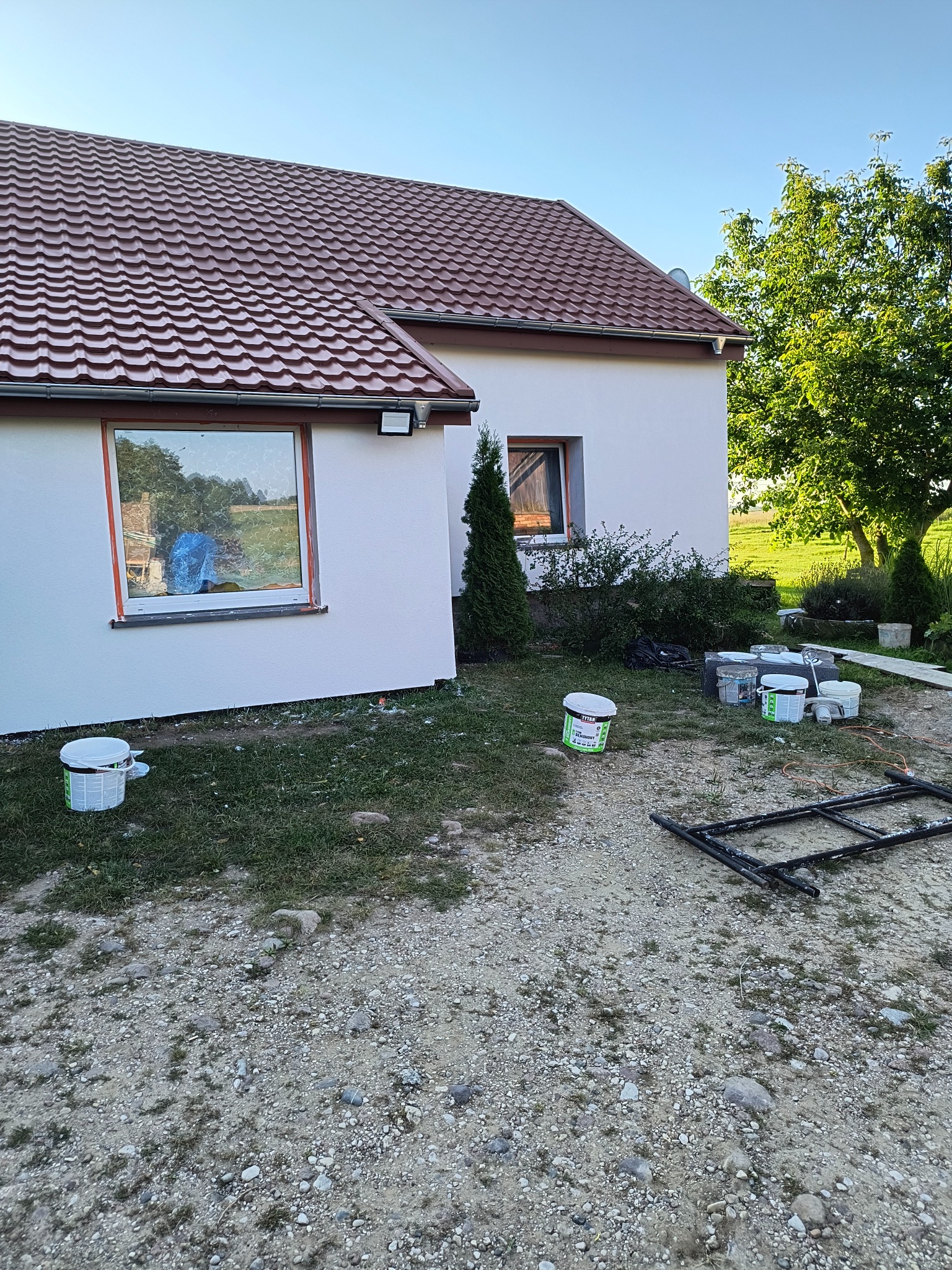 Biała elewacja domu z brązowym dachem po malowaniu, okna zabezpieczone folią. Na pierwszym planie puszki z farbą i rusztowanie.