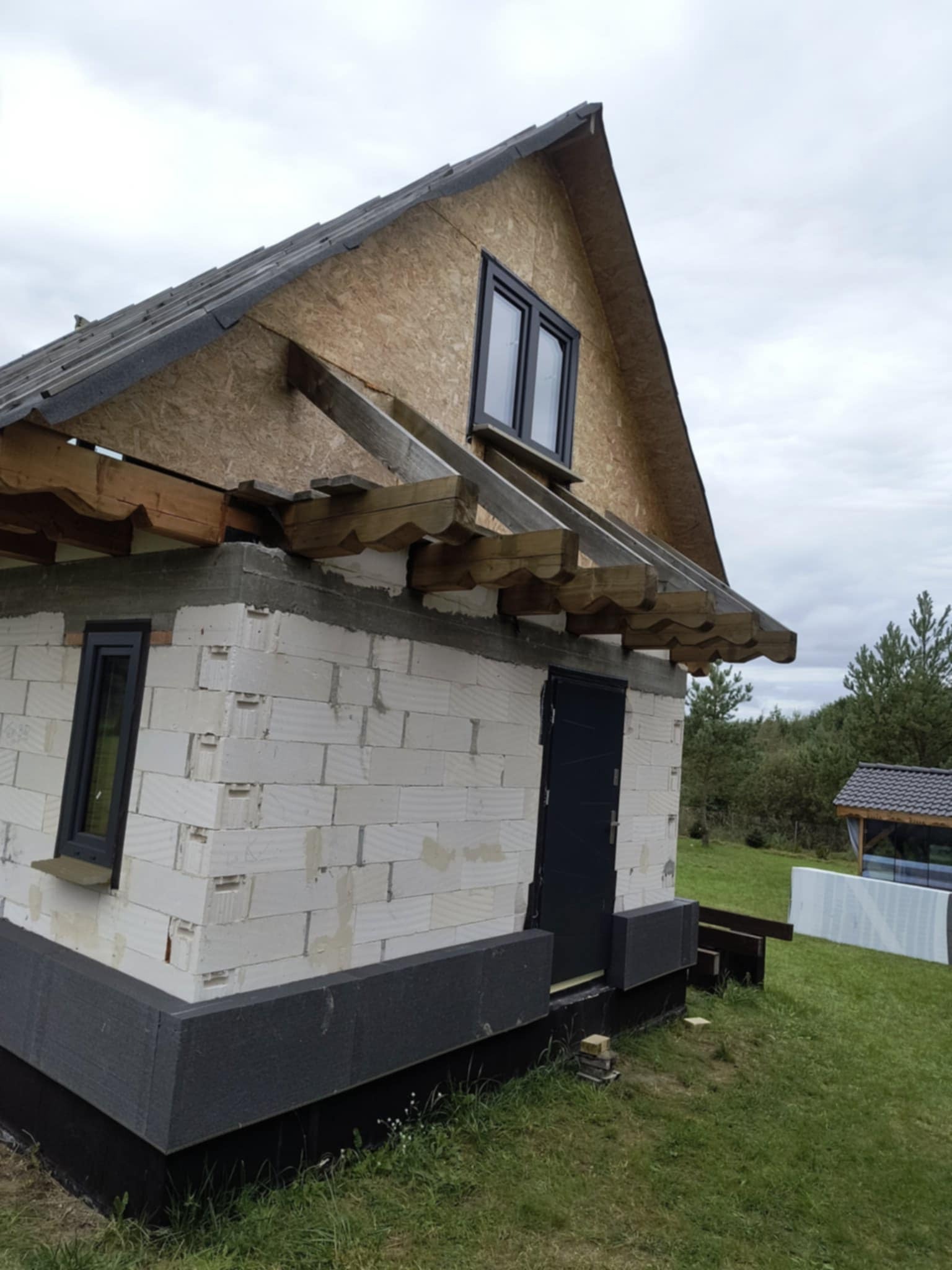 Budynek w trakcie budowy: białe bloczki, czarna stolarka okienna i drzwiowa, ocieplenie fundamentów, drewniana konstrukcja dachu z deskowaniem. Widok z zewnątrz, dzień.