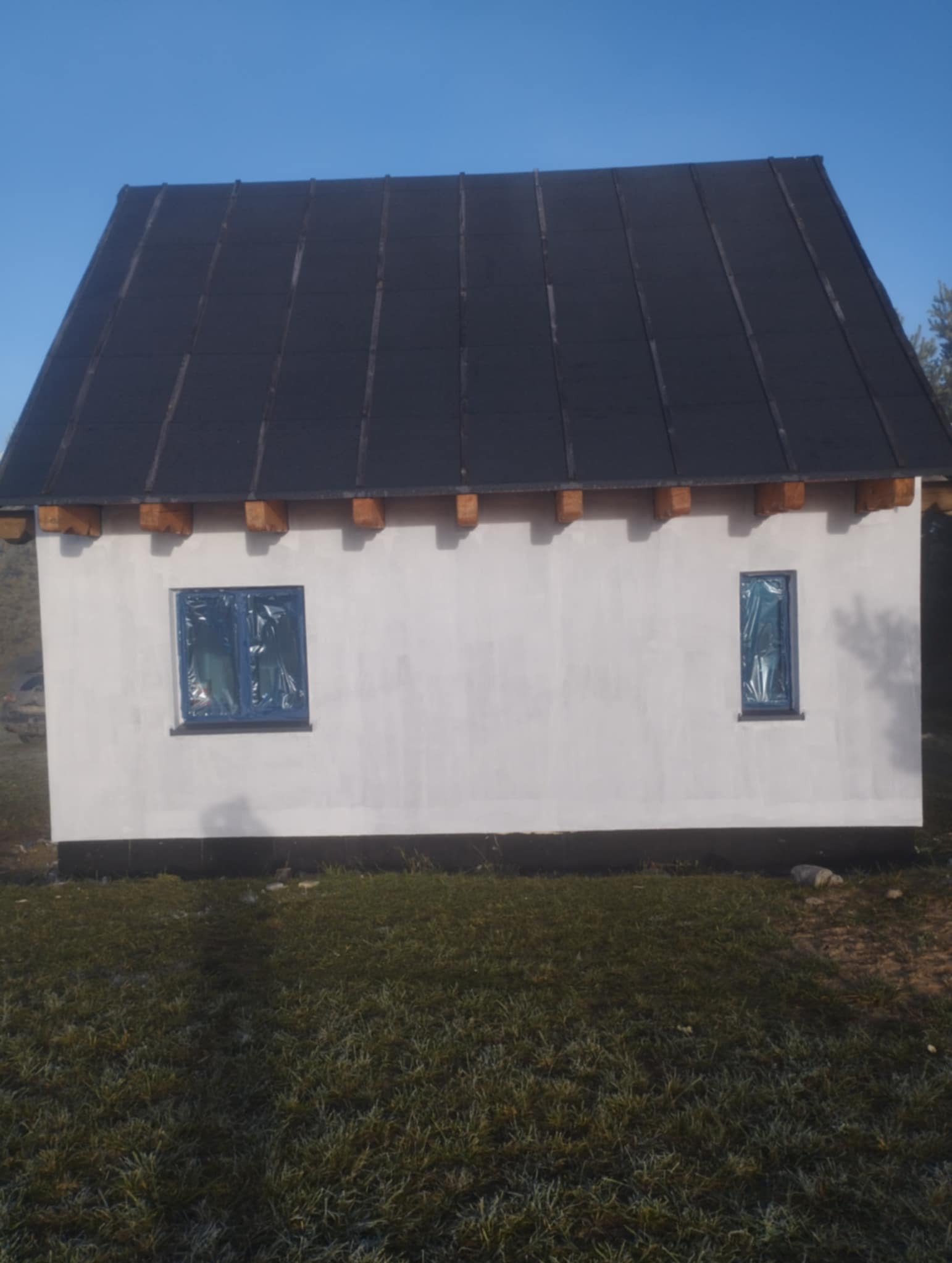 Niski dom z ciemnym dachem i otynkowaną, białą elewacją. Okna zabezpieczone folią. Widok z poziomu trawnika w słoneczny dzień.