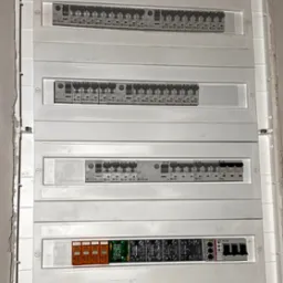 Rozdzielnica elektryczna w białej obudowie, zamontowana na ścianie w stanie surowym. Widoczne zabezpieczenia, bezpieczniki i moduły. Instalacja po montażu.