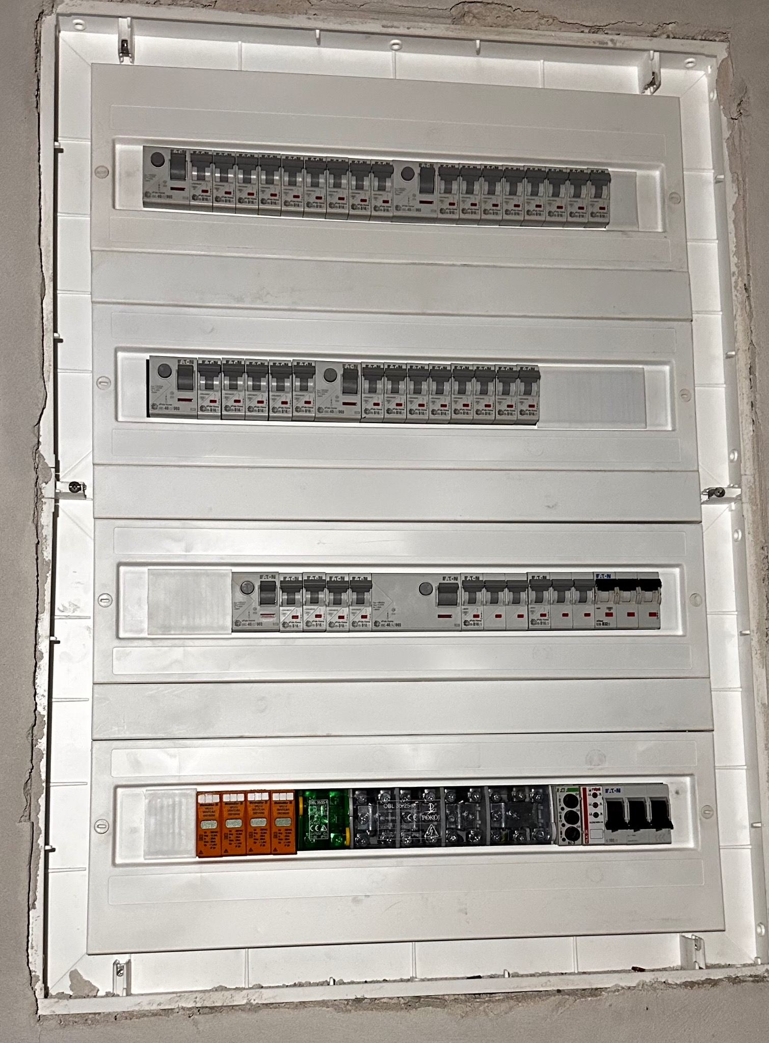 Rozdzielnica elektryczna w białej obudowie, zamontowana na ścianie w stanie surowym. Widoczne zabezpieczenia, bezpieczniki i moduły. Instalacja po montażu.