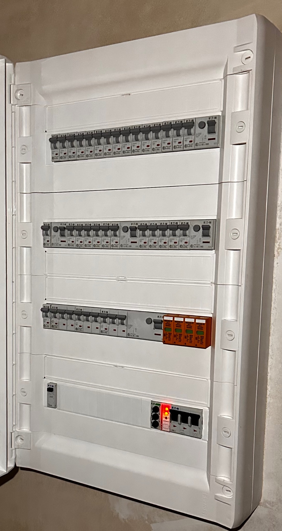 Otwarta skrzynka elektryczna z bezpiecznikami Eaton, widoczne trzy rzędy wyłączników i pomarańczowe zabezpieczenia. Dolny rząd z czerwonymi diodami sygnalizacyjnymi.