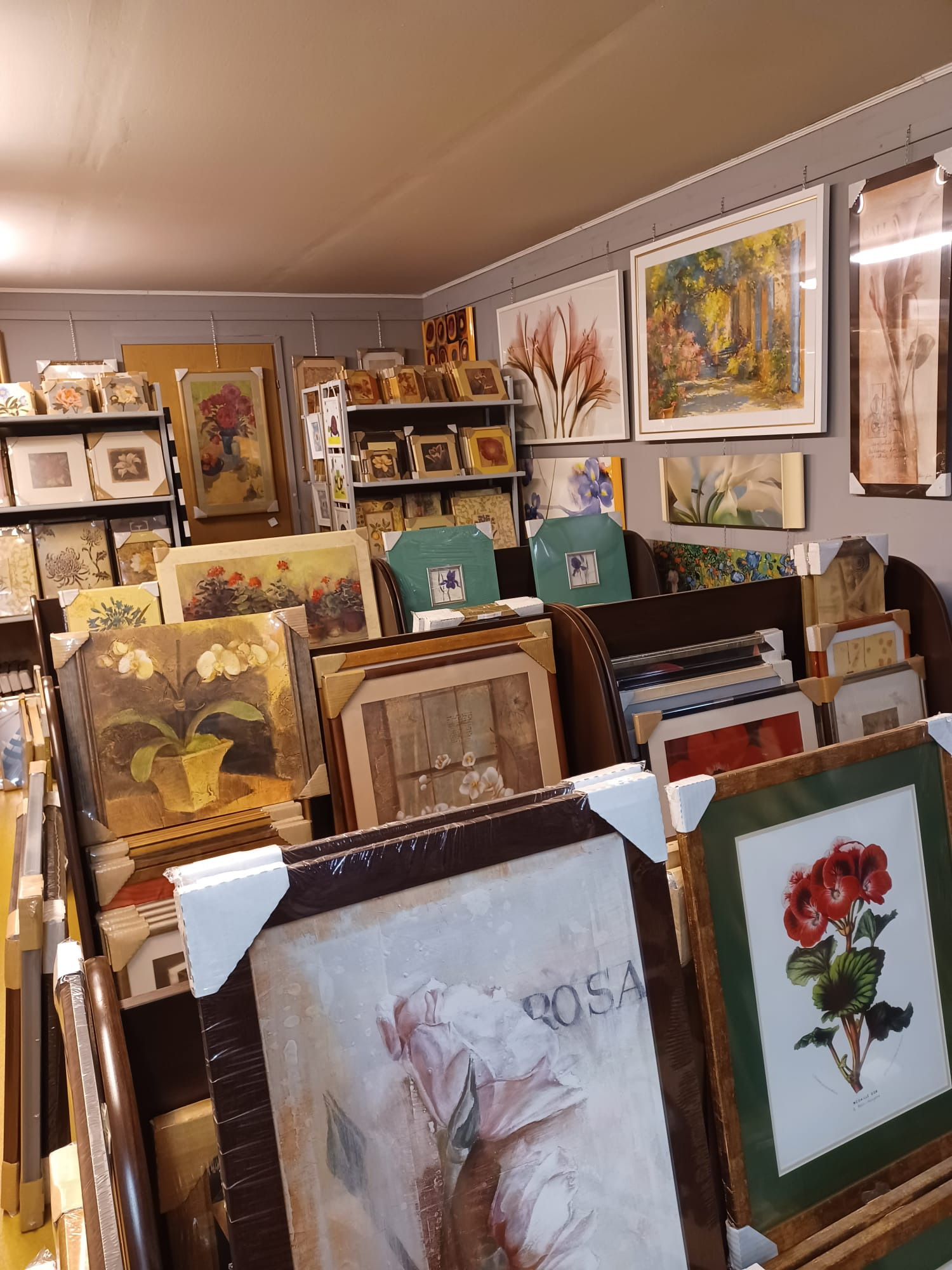 Sklep z obrazami: różnorodne, oprawione grafiki i obrazy z motywami kwiatowymi, ułożone na półkach i wiszące na ścianie, gotowe do zakupu.