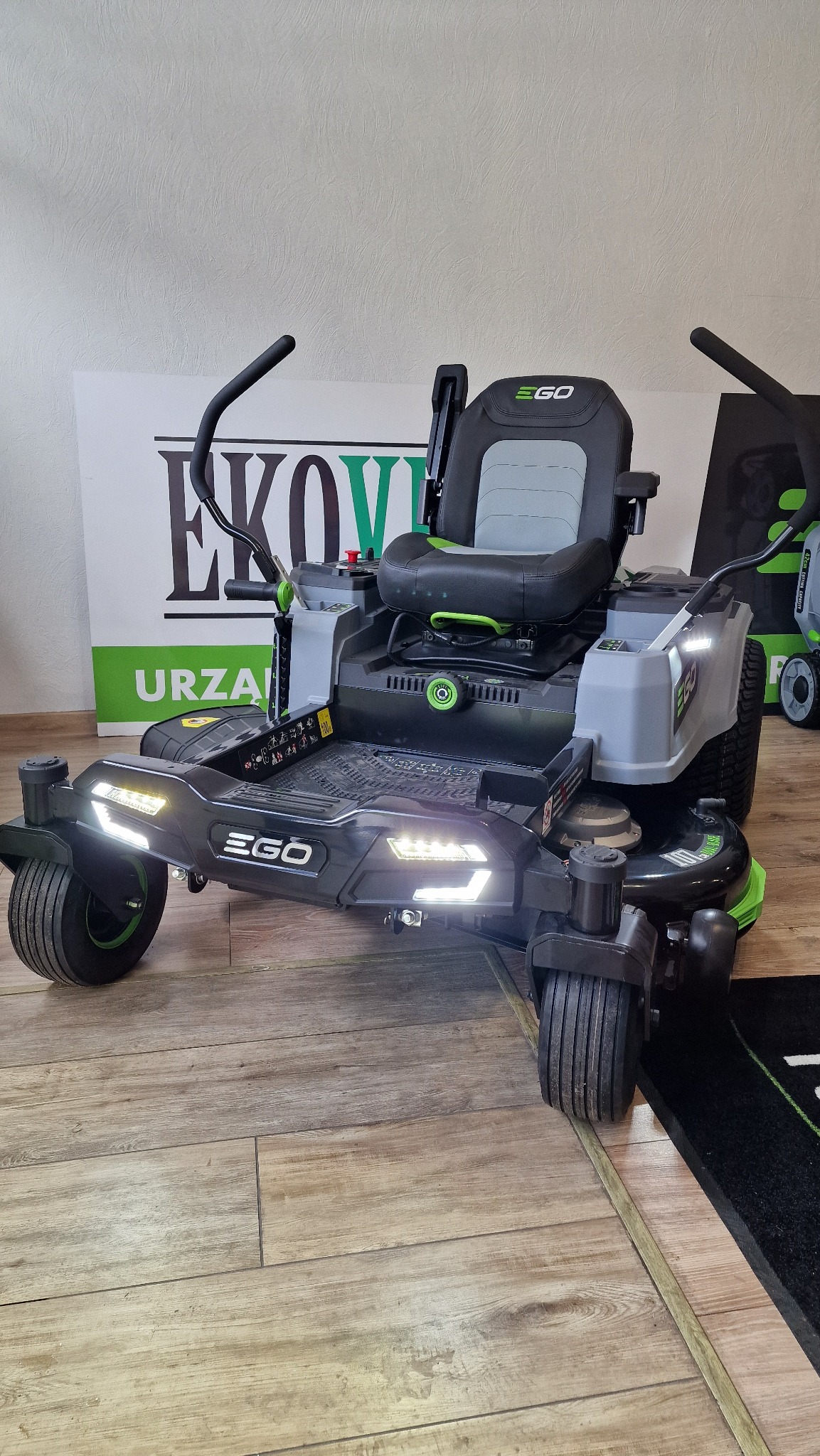 Elektryczna kosiarka samojezdna EGO z włączonymi światłami LED, prezentowana na drewnianej podłodze. Widoczny fragment ekspozycji z logo firmy.