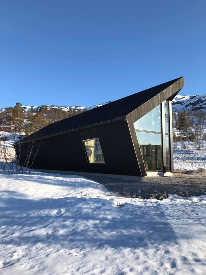 Nowoczesny, czarny dom szkieletowy z dużymi przeszkleniami, otoczony śnieżnym krajobrazem w słoneczny dzień. Nietypowy kształt dachu.