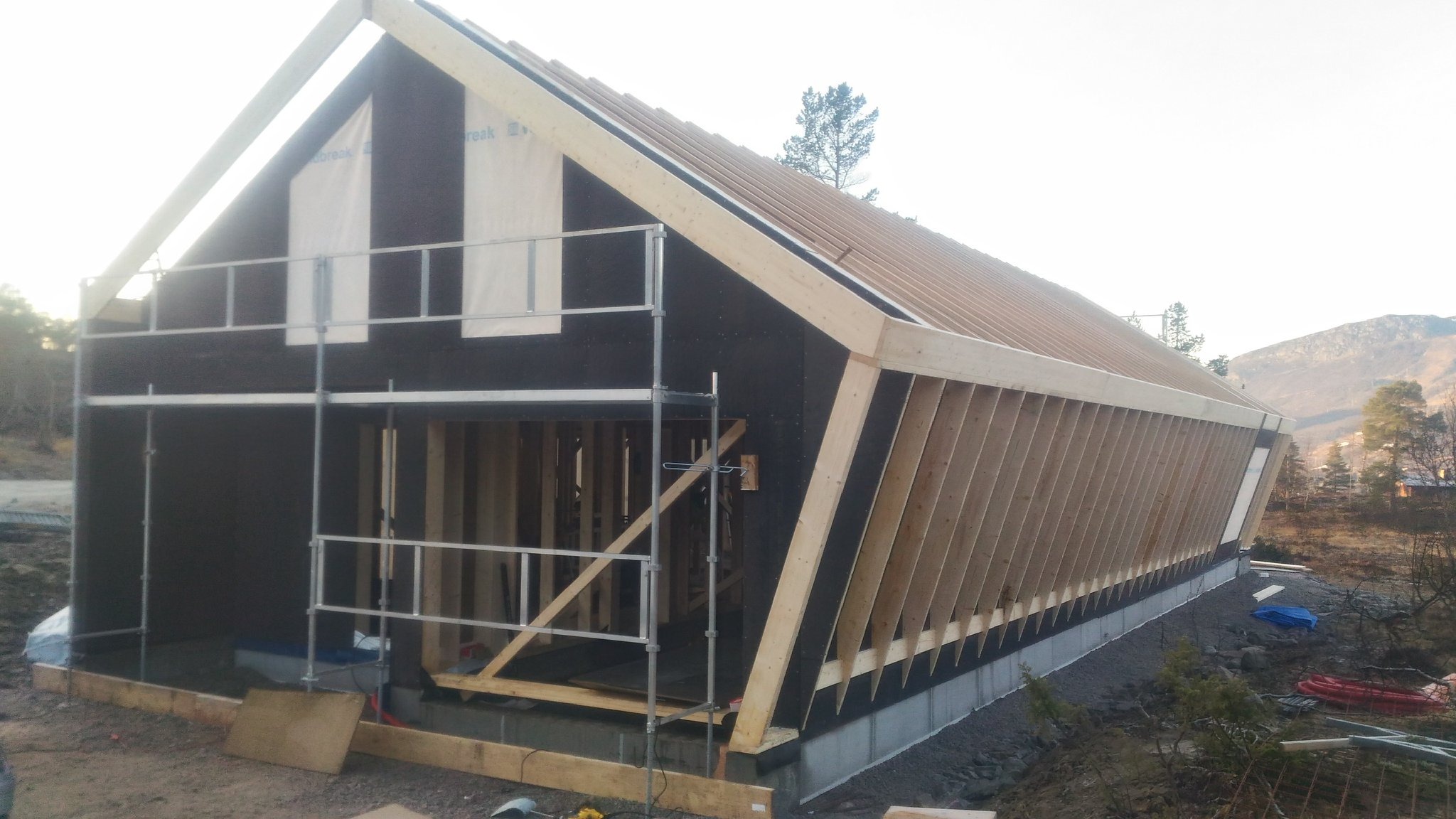 Szkielet domu w trakcie budowy, widoczne drewniane elementy konstrukcyjne, czarna membrana i rusztowanie. W tle górzysty krajobraz.