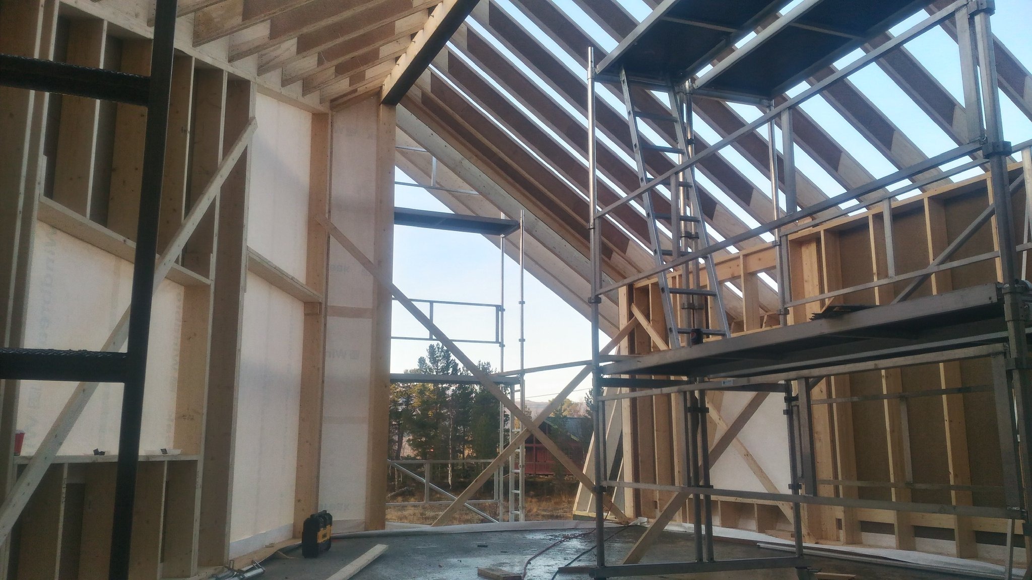 Wnętrze domu w budowie: drewniana konstrukcja szkieletowa z widocznym oknem, rusztowania i narzędzia na podłodze. Etap wznoszenia, widok na zewnątrz.