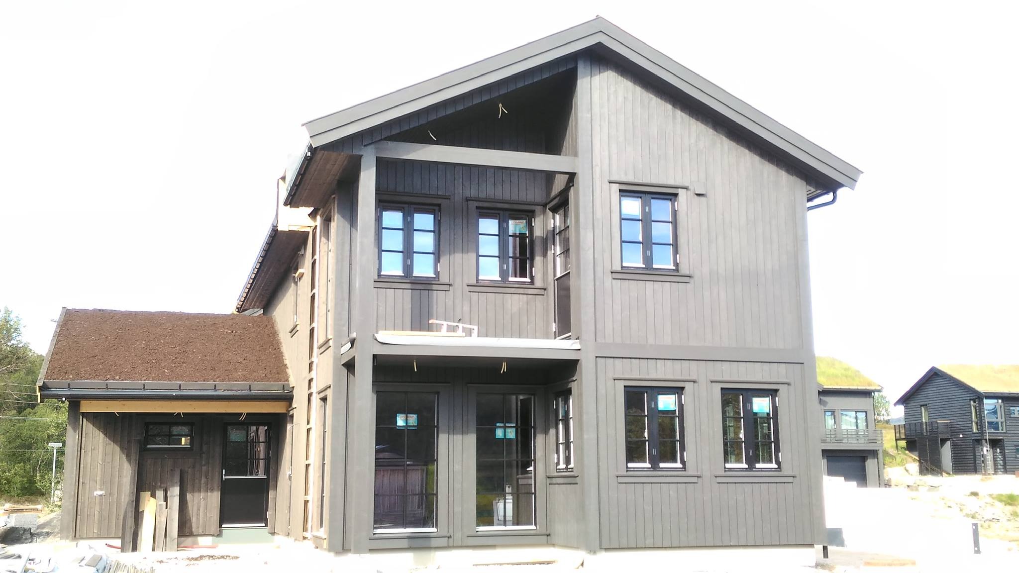 Nowoczesny dom szkieletowy z ciemną elewacją i oknami z czarnymi ramami. Widoczny fragment dachu pokrytego roślinnością. Dom dwukondygnacyjny.