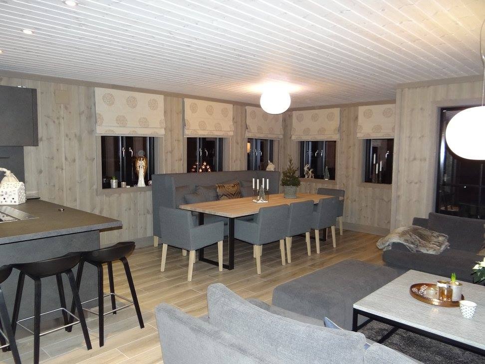 Wnętrze domu z drewnianymi ścianami i podłogą, szare meble, okna z roletami rzymskimi. Widoczna kuchnia z wyspą i jadalnia z narożną ławą.