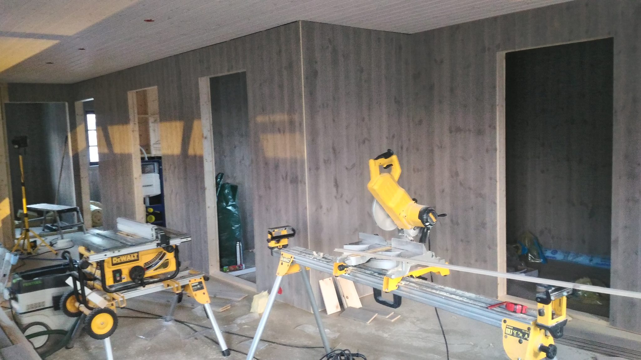 Wnętrze w trakcie wykończenia panelami ściennymi imitującymi drewno. Widoczne narzędzia: piły stołowe i ukośnice. Częściowo widoczne otwory drzwiowe.
