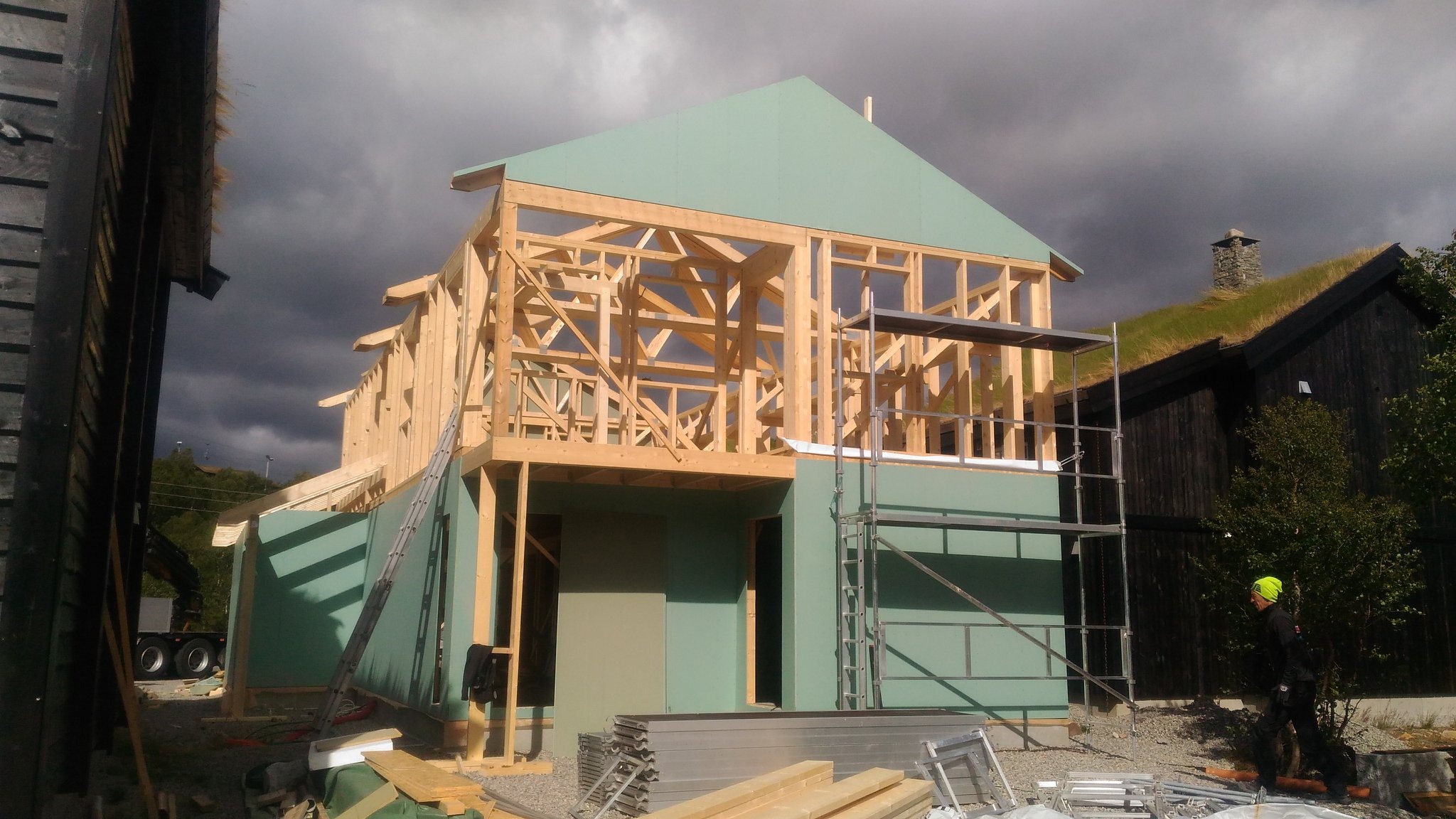 Konstrukcja szkieletowa domu w trakcie budowy z zielonymi płytami, rusztowaniem i stertą materiałów na pierwszym planie, na tle pochmurnego nieba.