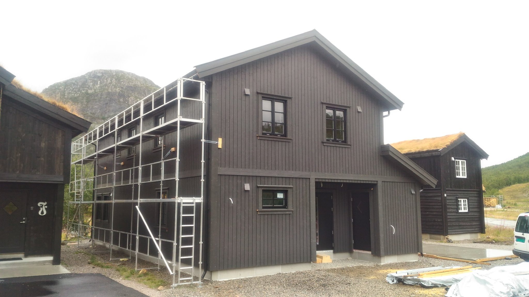 Ciemny, drewniany dom z rusztowaniem na tle gór. Budynek z dachem porośniętym trawą. Widoczny fragment drugiego budynku. Etap budowy/rozbudowy.