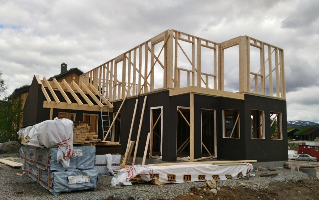 Wznoszenie domu szkieletowego: konstrukcja z drewnianych belek, czarne ściany, przygotowane materiały budowlane na placu budowy. Widok na góry w tle.
