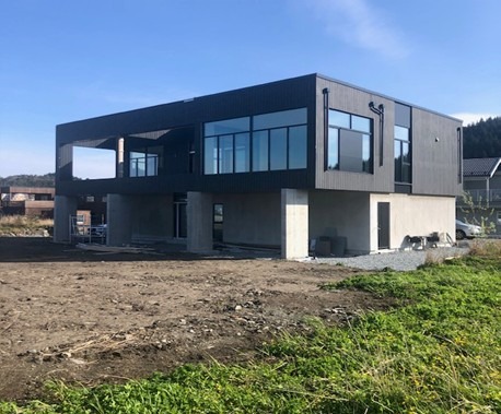 Nowoczesny, dwukondygnacyjny dom z czarną elewacją i dużymi oknami, wsparty na betonowych filarach, na tle zieleni i błękitnego nieba.