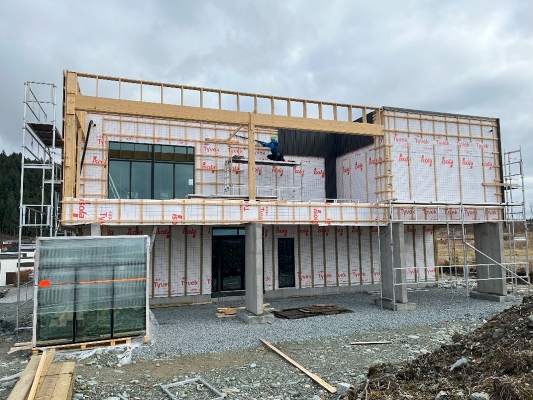 Budowa domu szkieletowego: montaż drewnianej konstrukcji, ocieplenie ścian folią izolacyjną, pracownik na rusztowaniu. Widoczne okna i materiały budowlane.