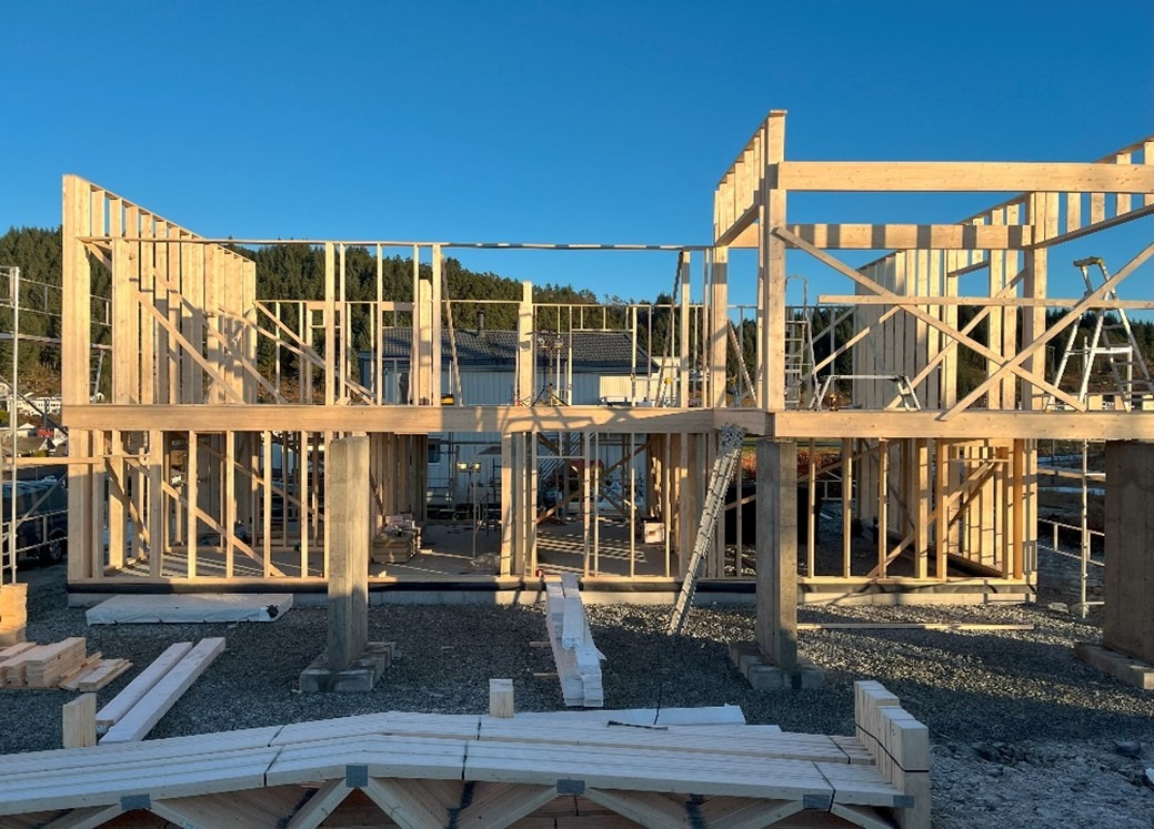 Konstrukcja szkieletowa domu w trakcie budowy, widok na dwie kondygnacje, drewniane belki, słupy, w tle budynki i drzewa, słoneczny dzień.