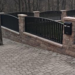 Ogrodzenia Kompleks - Czarne, metalowe ogrodzenie na niskim, kamiennym murku z czarną skrzynką pocztową, widoczne na tle drzew bez liści i szarej kostki brukowej.