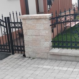 Ogrodzenia Kompleks - Otwarta, czarna, metalowa furtka z dekoracyjnym wzorem, osadzona między dwoma słupkami z jasnych, kamiennych bloczków. W tle fragment ogrodzenia i zielony trawnik.