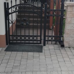 Ogrodzenia Kompleks - Czarne, metalowe ogrodzenie z bramką, zdobione ornamentami, oddziela chodnik z kostki od schodów prowadzących do domu. Widoczny fragment elewacji i zadaszenie.