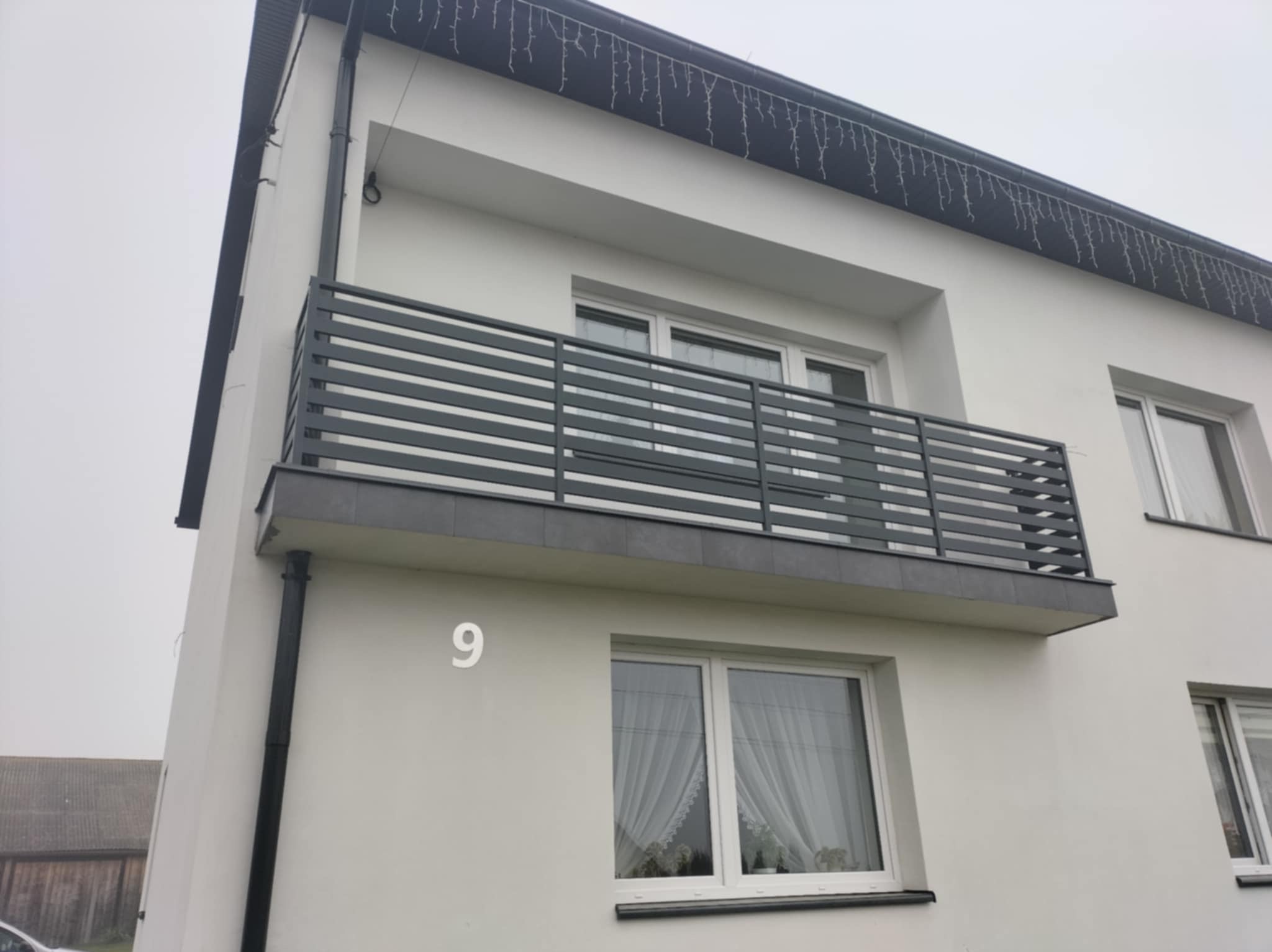Nowoczesna, pozioma balustrada balkonowa w kolorze grafitowym, zamontowana na białym budynku z numerem 9. Minimalistyczny design, szara podbitka dachu.