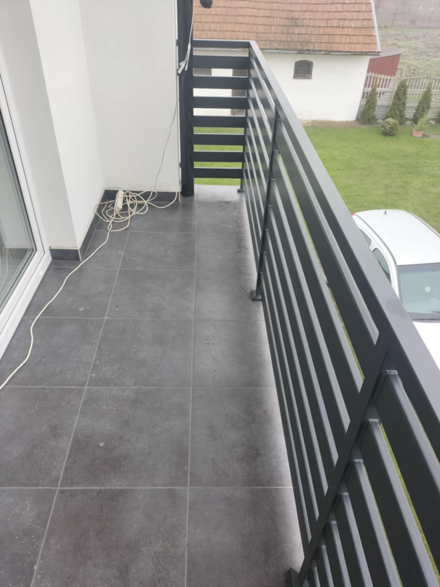 Nowoczesna, grafitowa balustrada balkonowa z poziomych profili. Widok z góry na szare płytki, fragment budynku i samochodu w tle, na zewnątrz.