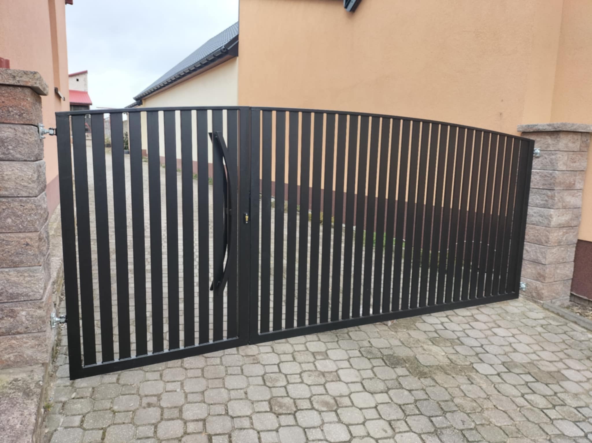 Czarna, metalowa brama posesyjna z pionowymi szczeblami, zamontowana między kamiennymi słupkami. Brama na tle domu z beżową elewacją i szarą kostką brukową.