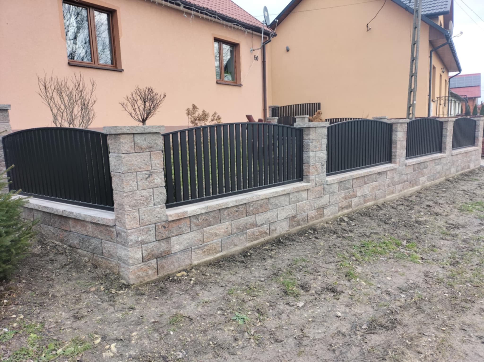 Czarno-szare ogrodzenie metalowe z podmurówką z bloczków betonowych, widok z zewnątrz posesji, na tle dwukondygnacyjnego domu w beżowej kolorystyce.