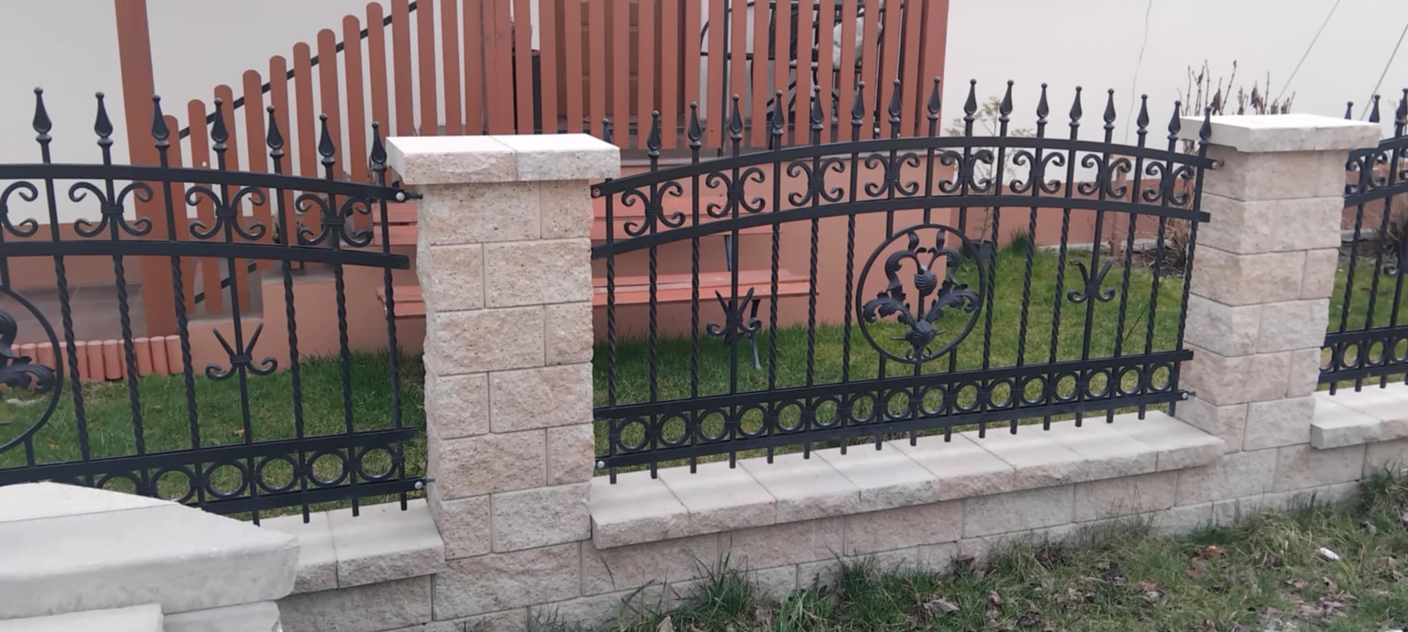 Czarne, kute ogrodzenie z kamiennymi słupkami i dekoracyjnymi elementami na tle zielonego trawnika i drewnianego płotu.