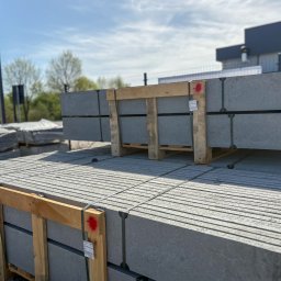 WYRGLAS Ogrodzenia Zbigniew Grasza - Szare, betonowe elementy ogrodzeniowe ułożone na paletach, zabezpieczone taśmami. Widoczne tło z budynkiem i drzewami. Materiały przygotowane do montażu.