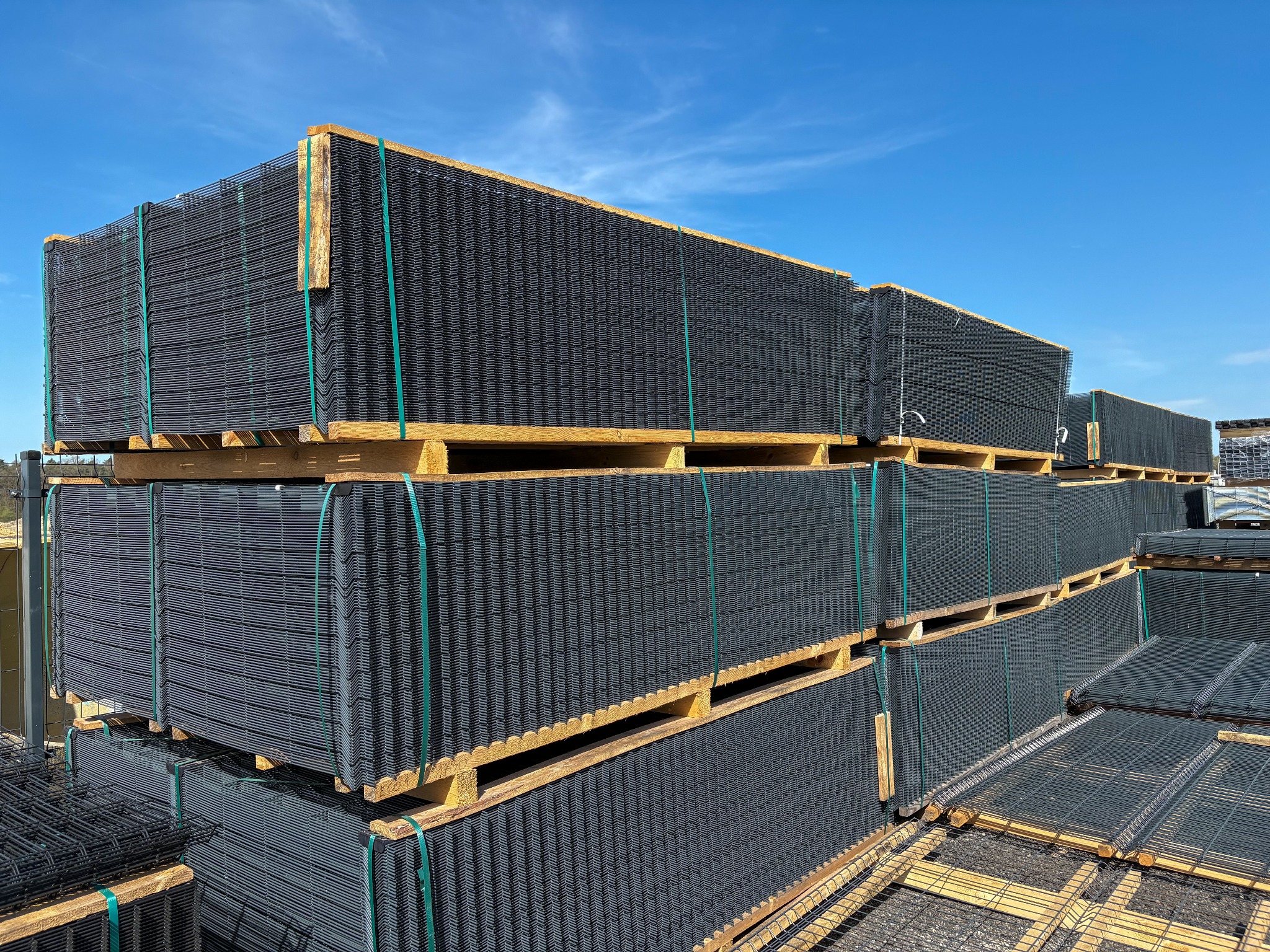 Stosy paneli ogrodzeniowych zgrzewanych, czarnych, zabezpieczonych zielonymi taśmami, ułożone na paletach pod błękitnym niebem. Widok z dołu.