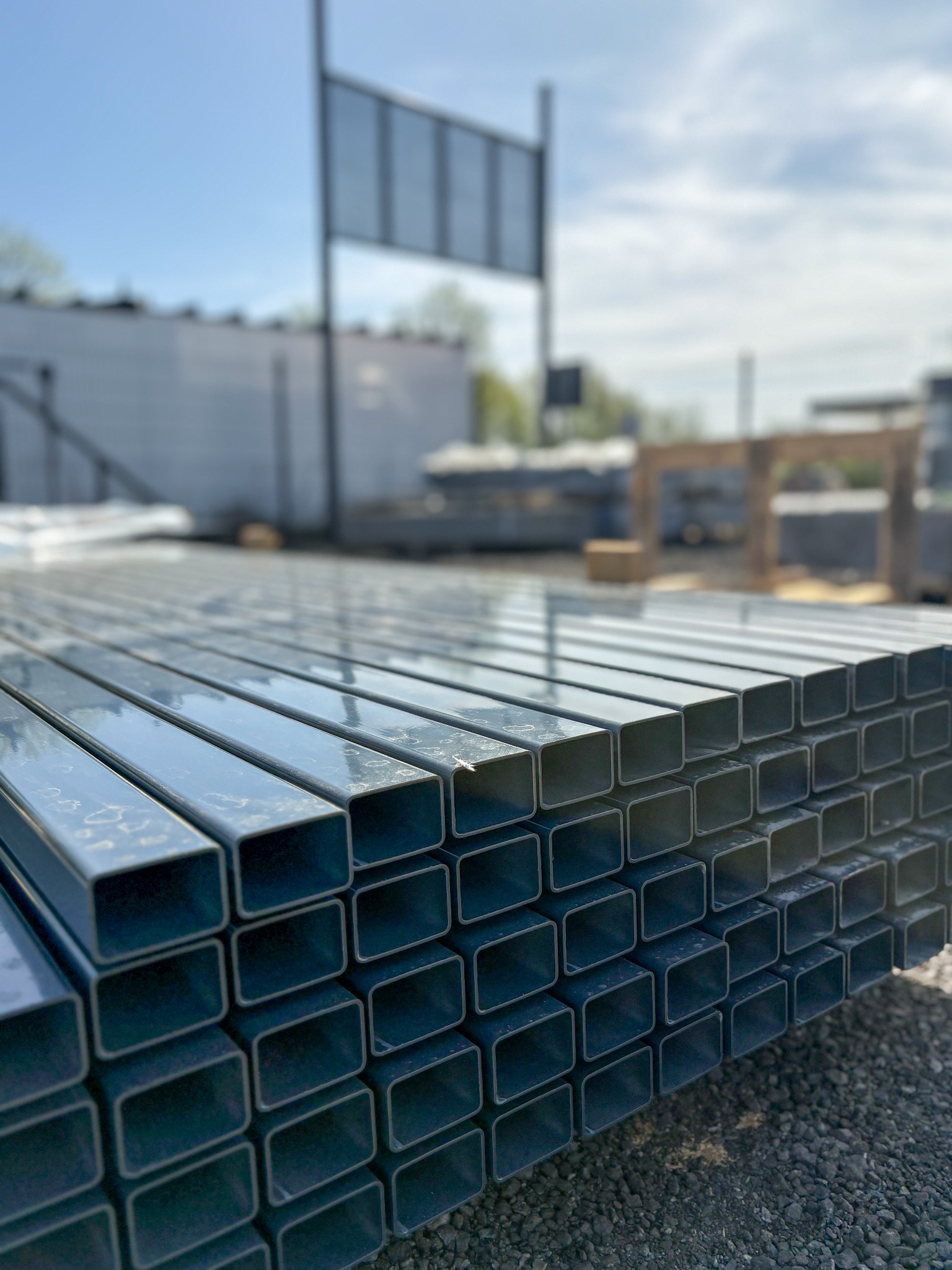 Stos ocynkowanych profili zamkniętych o kwadratowym przekroju, ułożonych na żwirowym podłożu, w tle magazyn i elementy konstrukcyjne.