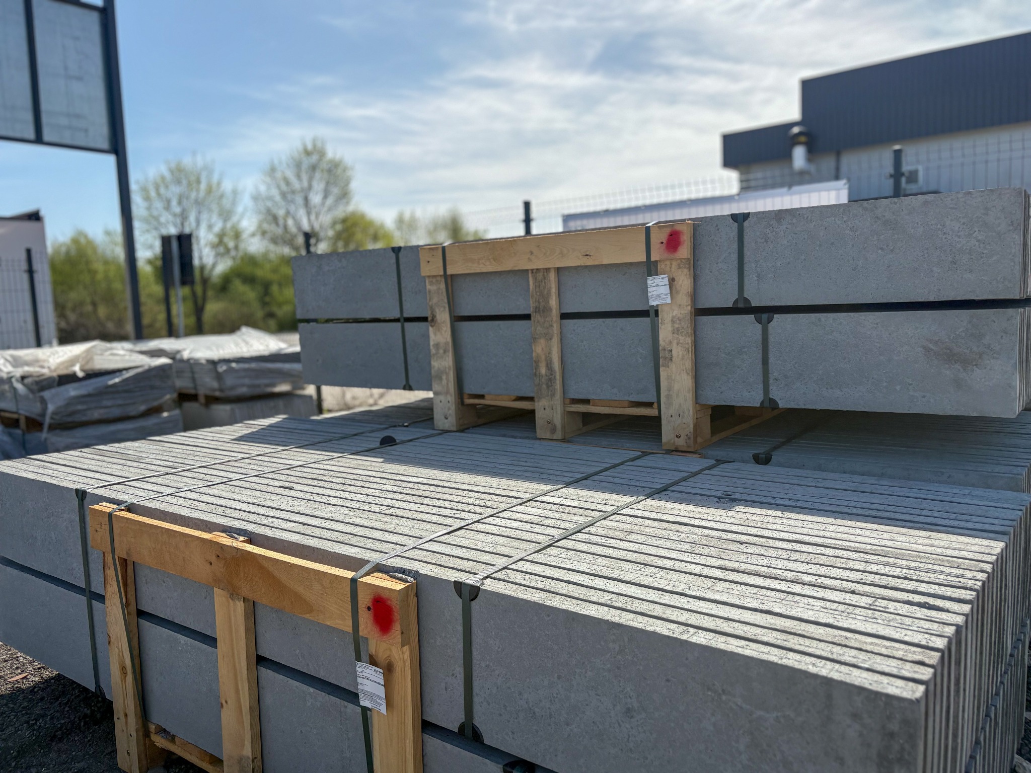 Szare, betonowe elementy ogrodzeniowe ułożone na paletach, zabezpieczone taśmami. Widoczne tło z budynkiem i drzewami. Materiały przygotowane do montażu.