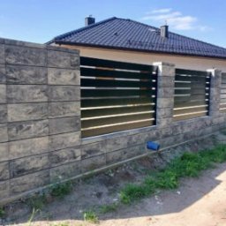 Towarzystwo Handlowe ALPLAST Sp. z o.o. Sp.k. - Nowoczesne ogrodzenie panelowe z elementami betonowymi w odcieniach szarości, z widocznym fragmentem domu w tle. Solidna konstrukcja, poziome panele.