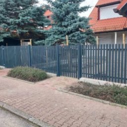 Towarzystwo Handlowe ALPLAST Sp. z o.o. Sp.k. - Grafitowe ogrodzenie panelowe z furtką, widok z chodnika na tle domu z czerwonym dachem i zielonymi drzewami. Wzdłuż ogrodzenia rosną krzewy.