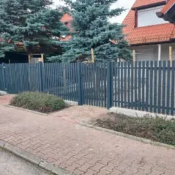 Grafitowe ogrodzenie panelowe z furtką, widok z chodnika na tle domu z czerwonym dachem i zielonymi drzewami. Wzdłuż ogrodzenia rosną krzewy.