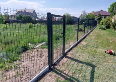 Zamontowane ogrodzenie panelowe z bramką na posesji, widok z boku, trawa, dom w tle, jasne niebo. Elementy betonowe u podstawy słupków.