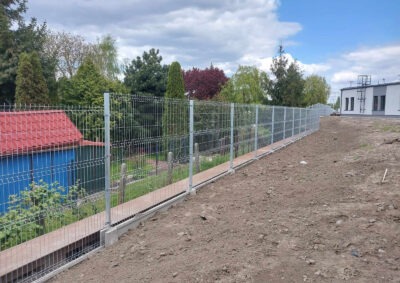 Długie, panelowe ogrodzenie z metalowej siatki, osadzone na betonowej podmurówce, oddzielające posesję z domem od terenu z budynkiem. Widok z boku, w tle drzewa.
