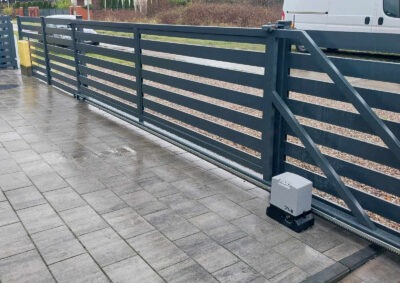 Automatyczna brama przesuwna z poziomymi szczeblami, szara, z widocznym mechanizmem napędowym na szarej kostce brukowej. W tle biały samochód dostawczy.